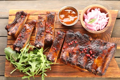 Photo of Delicious pork ribs, arugula, sauce and onion on wooden table, top view