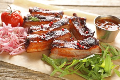 Photo of Delicious pork ribs, sauce, arugula and onion on wooden table, closeup