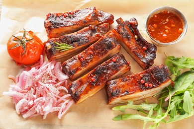 Photo of Delicious pork ribs, sauce, arugula and onion on parchment paper, top view