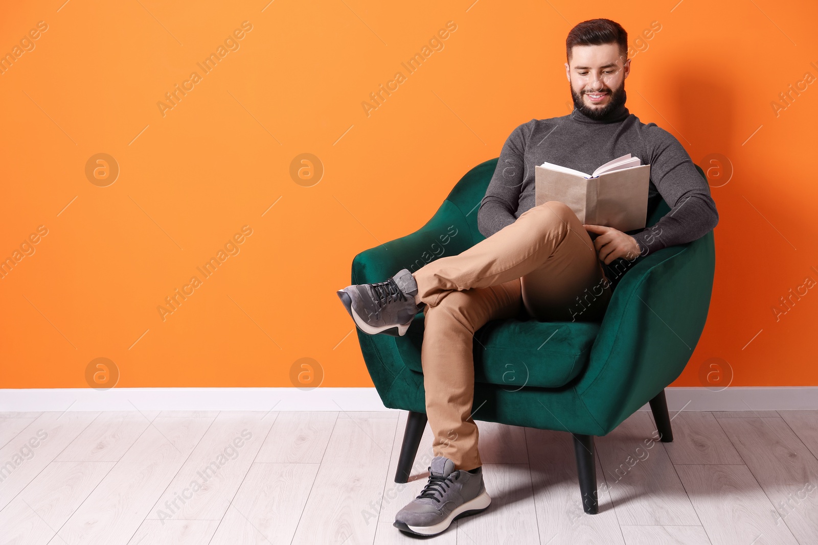 Smiling man with book relaxing on armchair near orange wall indoors. Space for text Photo of Smiling man with book relaxing on armchair near orange wall indoors. Space for text