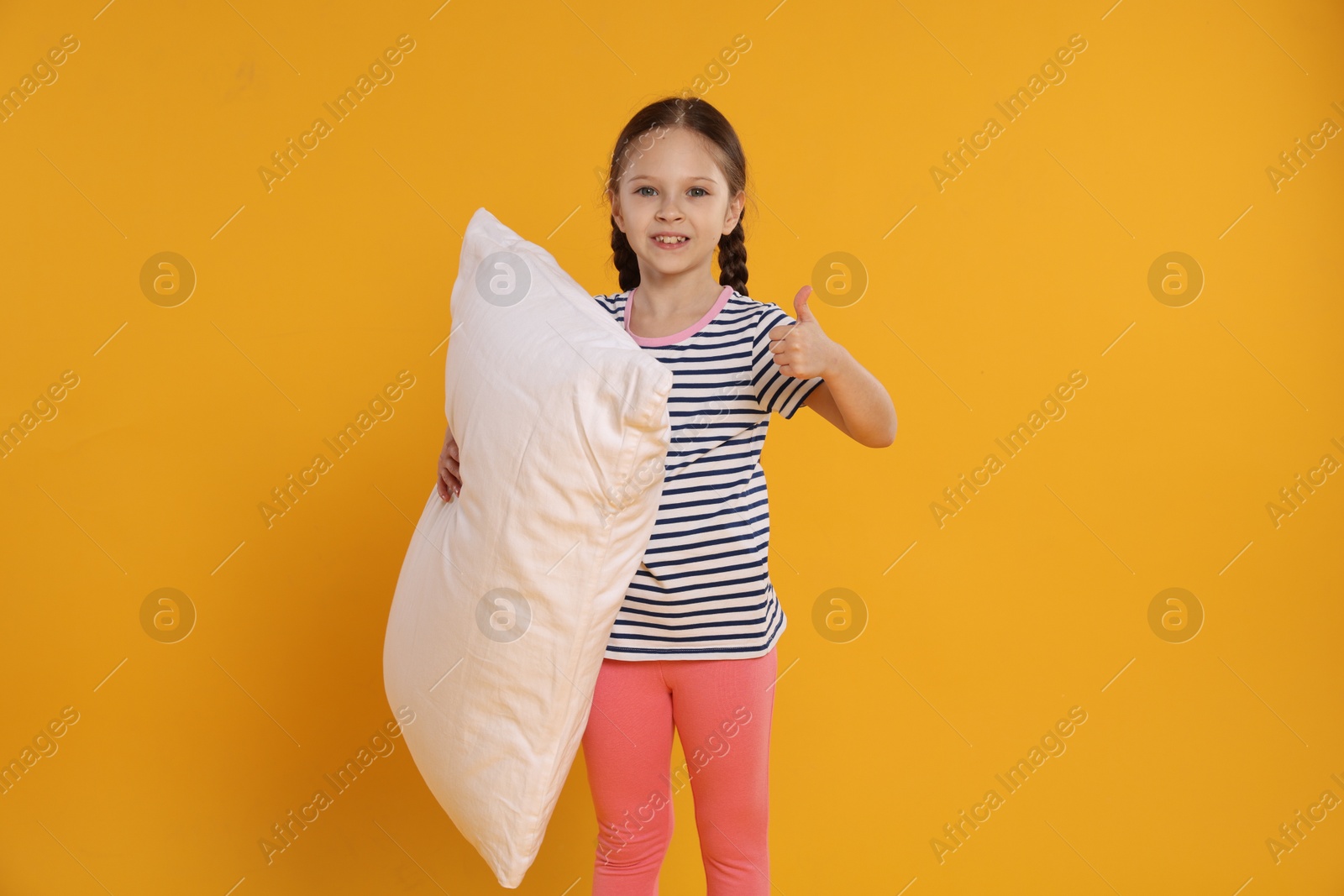 Cute girl with pillow showing thumbs up on orange background Photo of Cute girl with pillow showing thumbs up on orange background