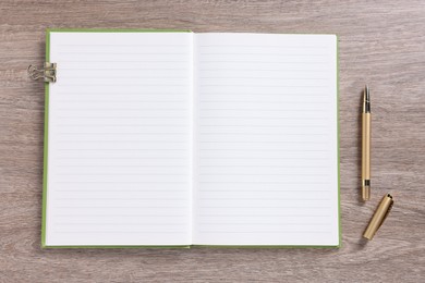 Photo of Open blank notebook, binder clip and pen on wooden table, top view