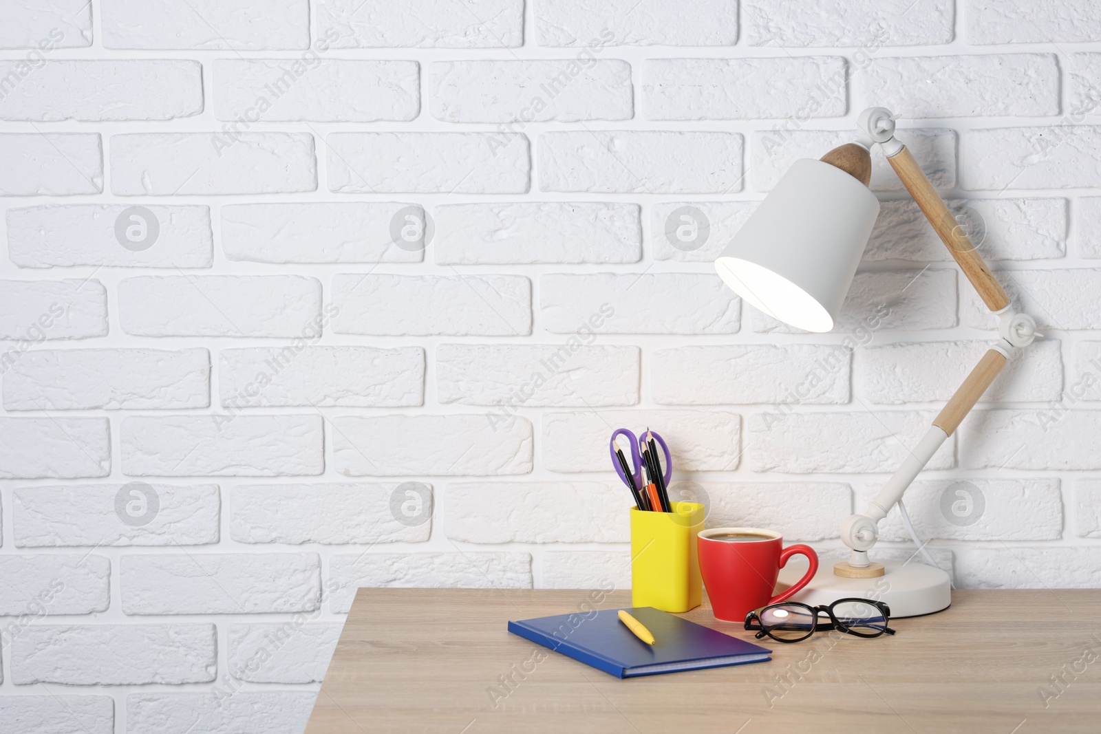 Different stationery, coffee, lamp and glasses on wooden office table near white brick wall, space for text Photo of Different stationery, coffee, lamp and glasses on wooden office table near white brick wall, space for text