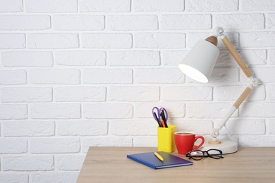 Photo of Different stationery, coffee, lamp and glasses on wooden office table near white brick wall, space for text
