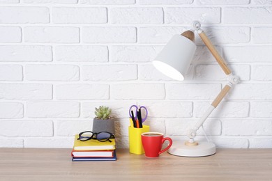 Photo of Different stationery, coffee, lamp, glasses and floral decor on wooden office table near white brick wall