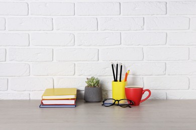 Photo of Different stationery, coffee, glasses and floral decor on wooden office table near white brick wall
