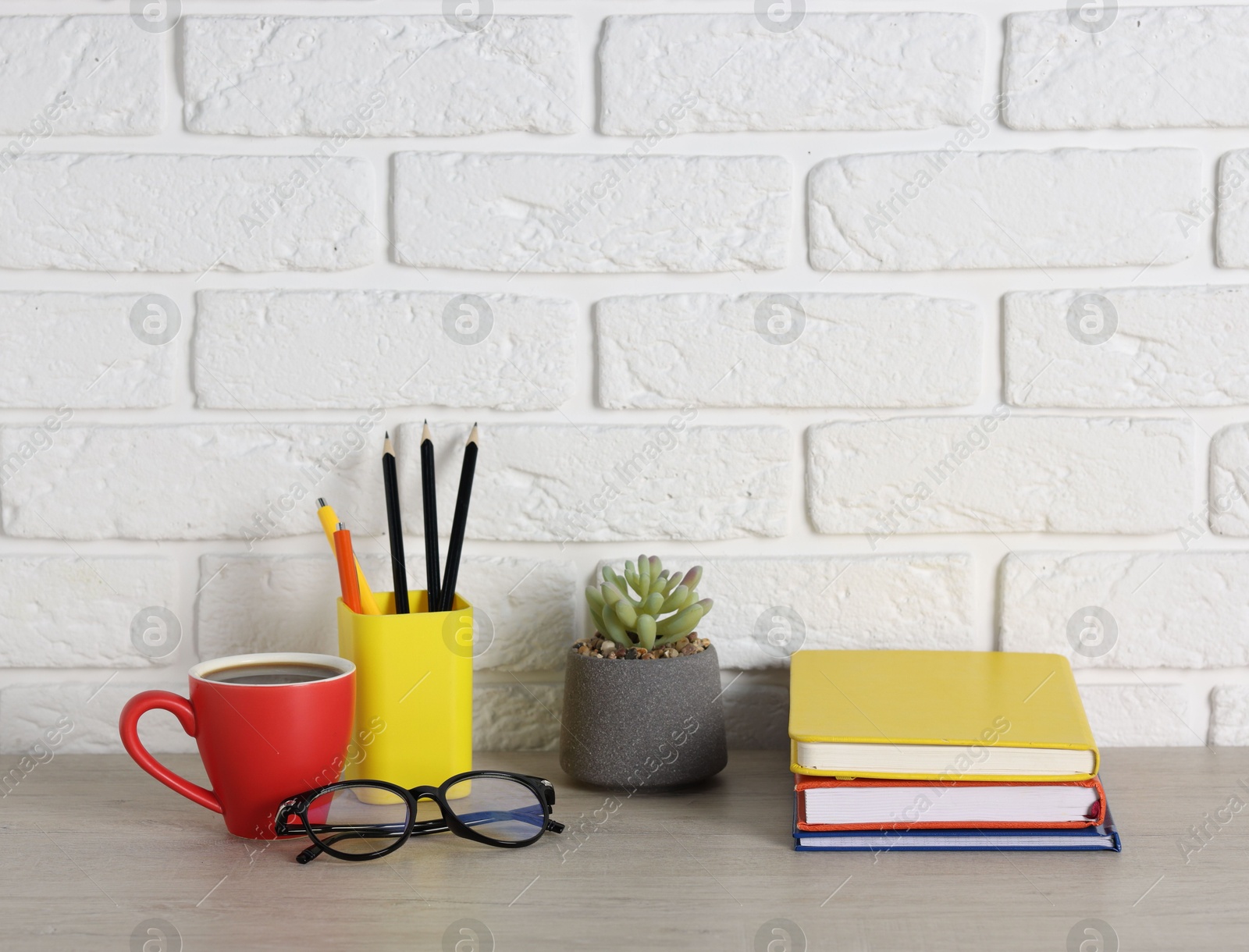 Different stationery, coffee, glasses and floral decor on wooden office table near white brick wall Photo of Different stationery, coffee, glasses and floral decor on wooden office table near white brick wall