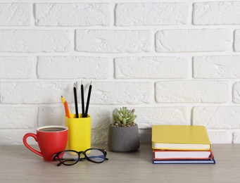 Photo of Different stationery, coffee, glasses and floral decor on wooden office table near white brick wall