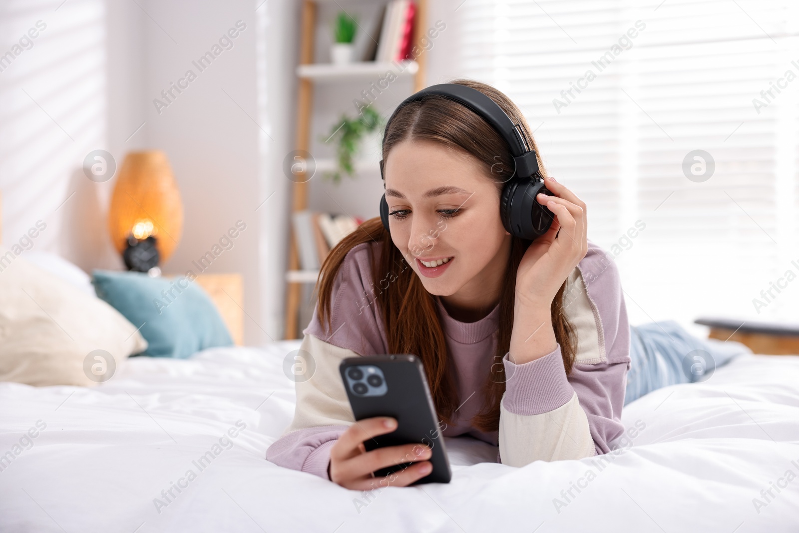Teenage girl in headphones using smartphone on bed at home, space for text Photo of Teenage girl in headphones using smartphone on bed at home, space for text