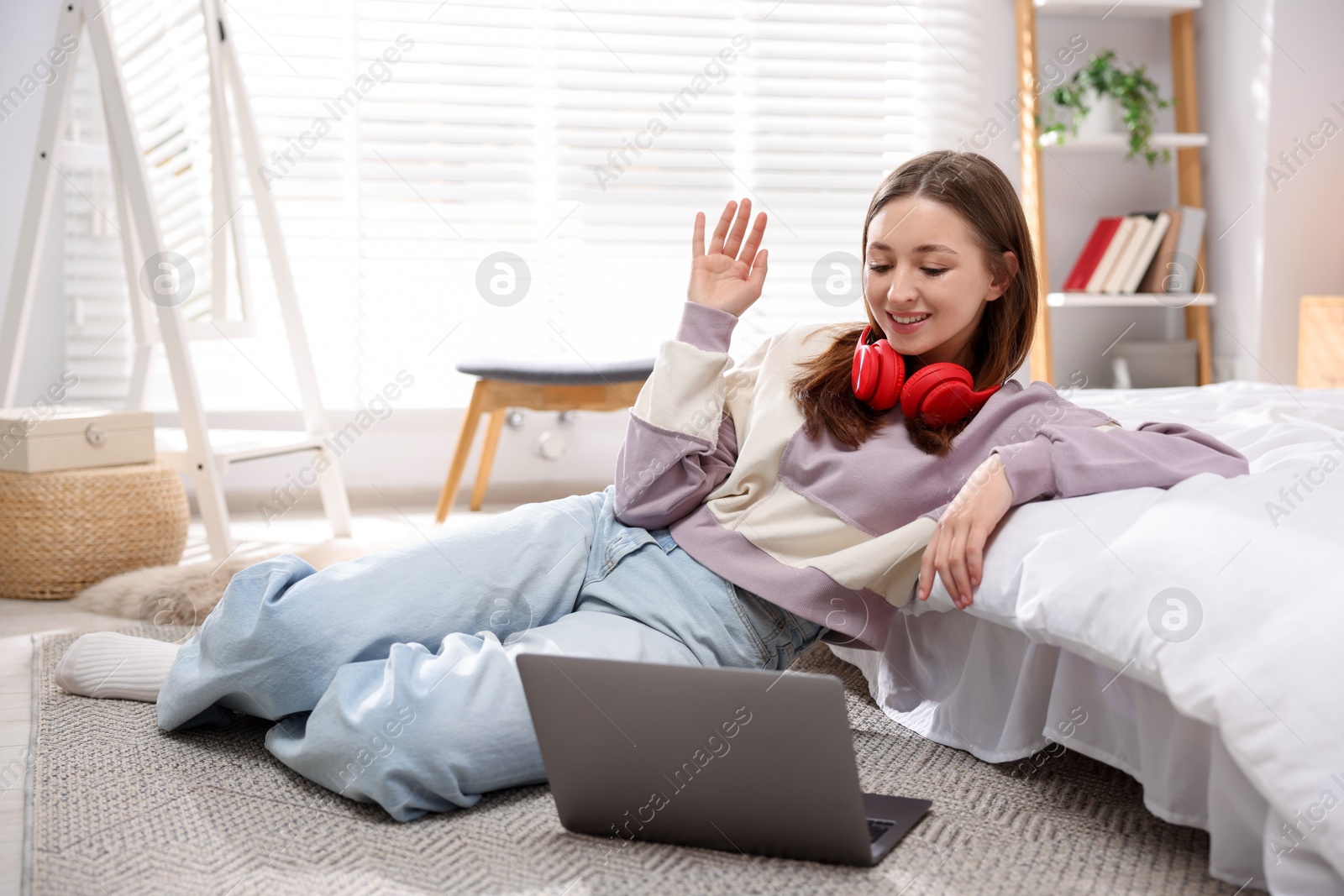Photo of Teenage girl with headphones having video chat via laptop on floor at home