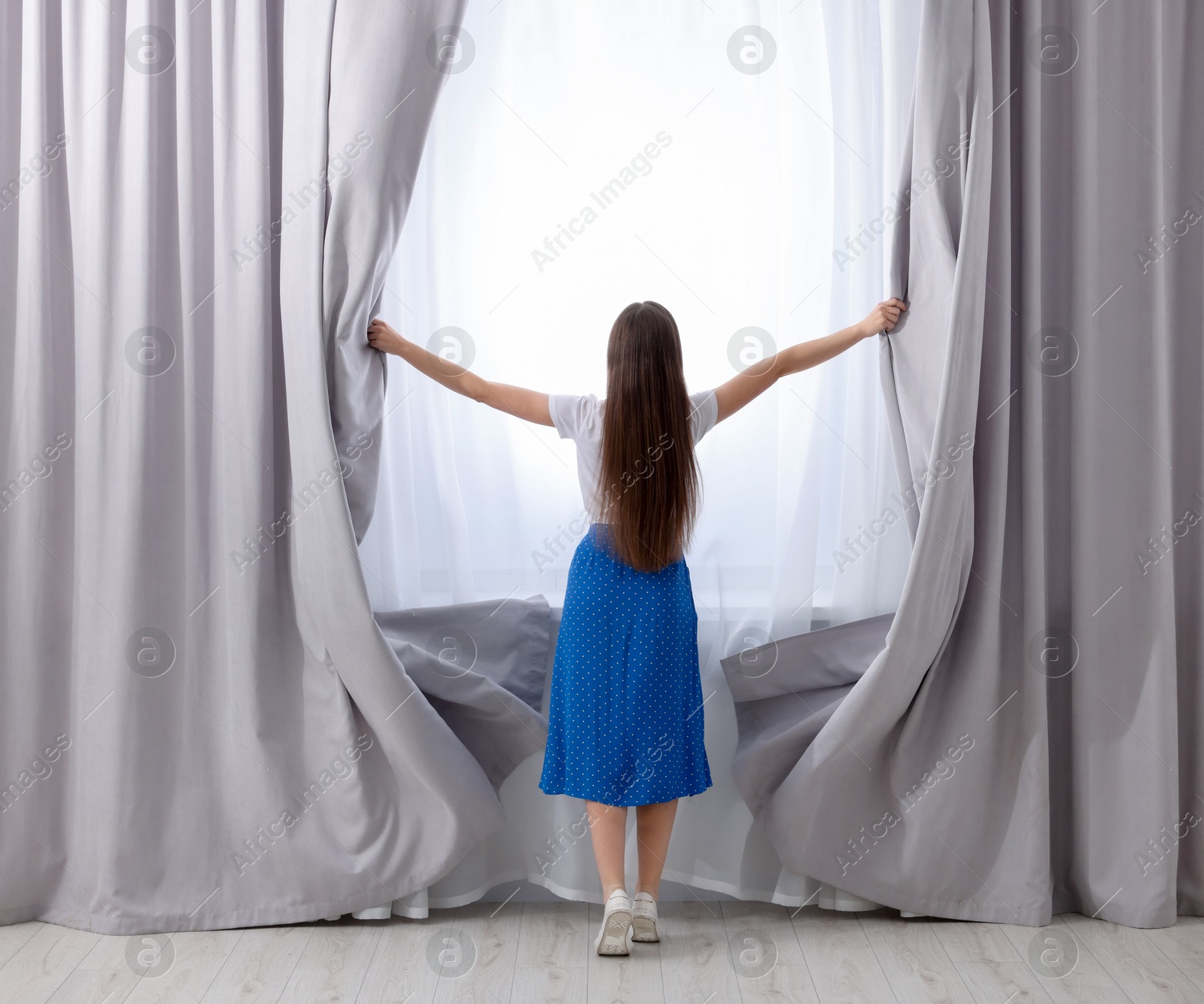 Woman opening stylish curtains at home, back view Photo of Woman opening stylish curtains at home, back view
