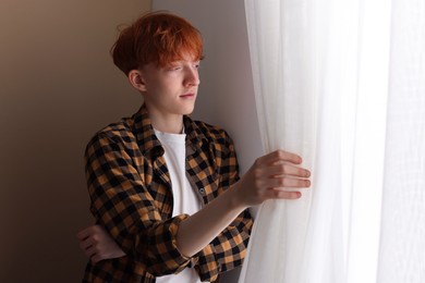 Photo of Teen problems. Depressed teenage boy looking out window indoors