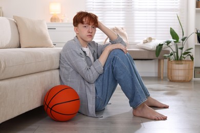 Photo of Teen problems. Depressed teenage boy with basketball ball on floor indoors