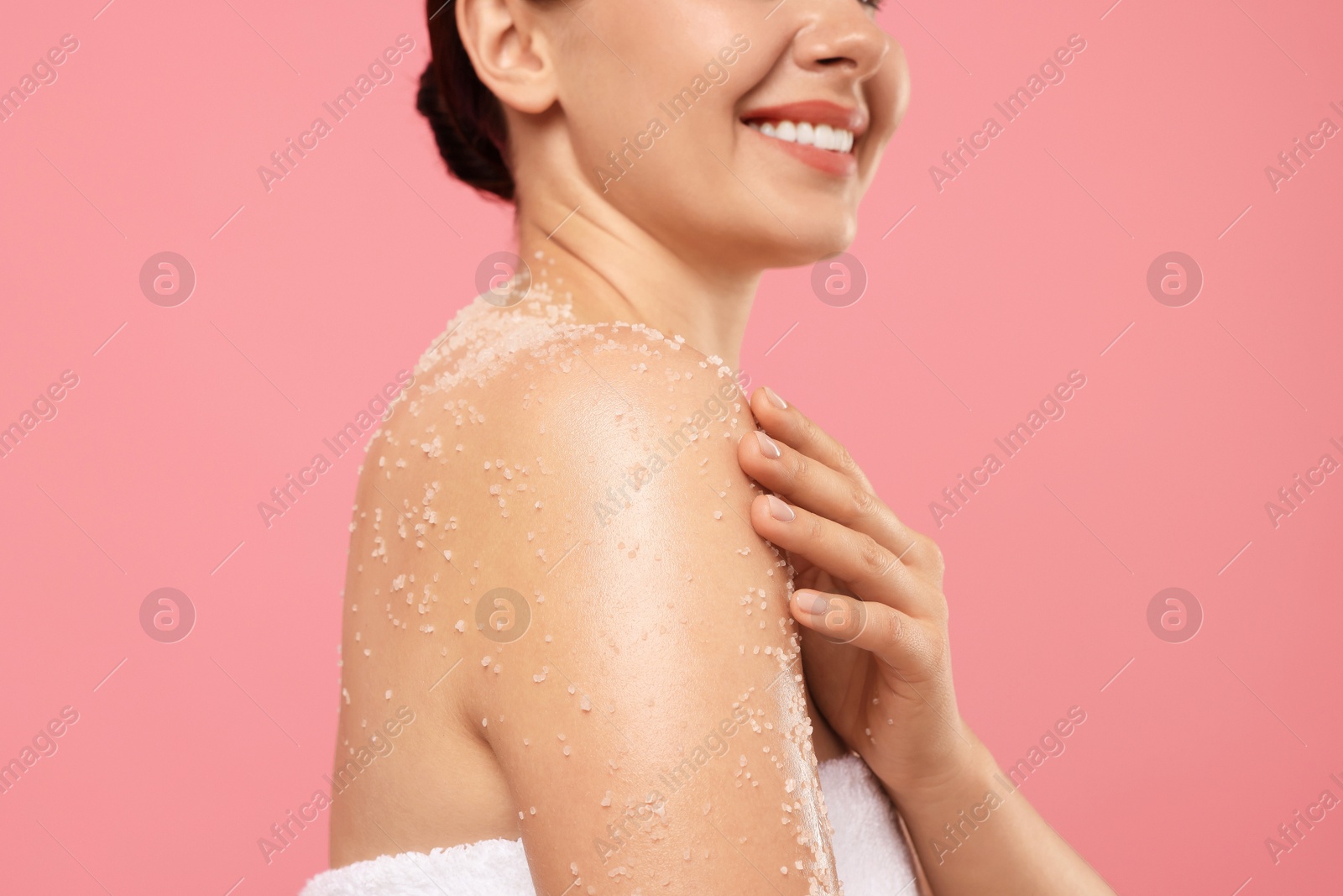 Photo of Smiling woman applying body scrub onto her shoulder against pink background, closeup