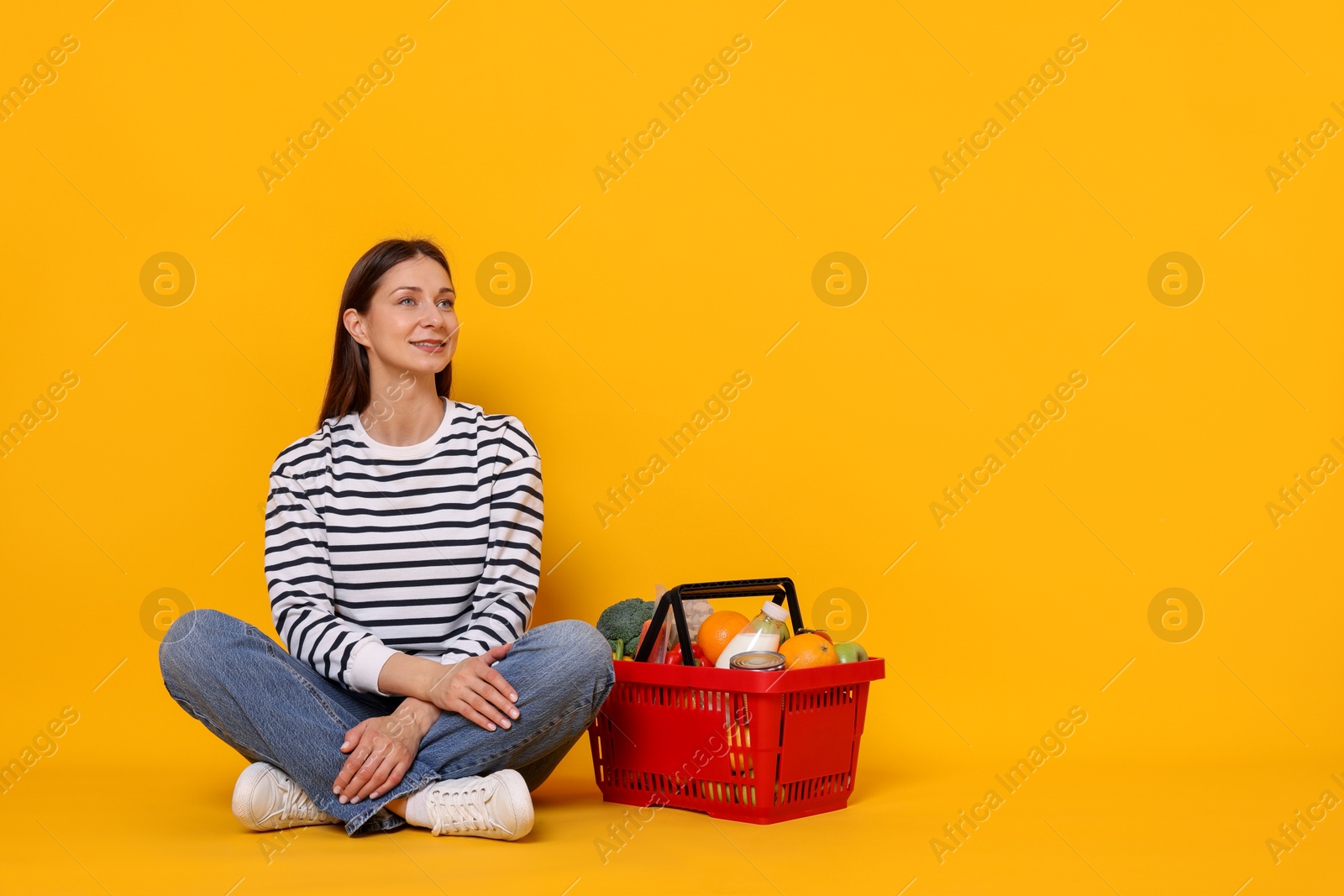 Smiling woman and shopping basket full of products on yellow background. Space for text Photo of Smiling woman and shopping basket full of products on yellow background. Space for text