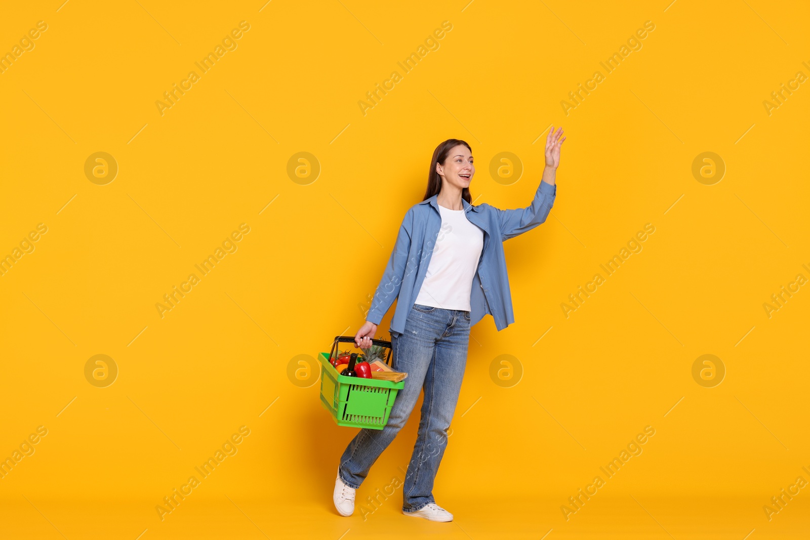 Smiling woman with shopping basket full of products waving hello on yellow background Photo of Smiling woman with shopping basket full of products waving hello on yellow background
