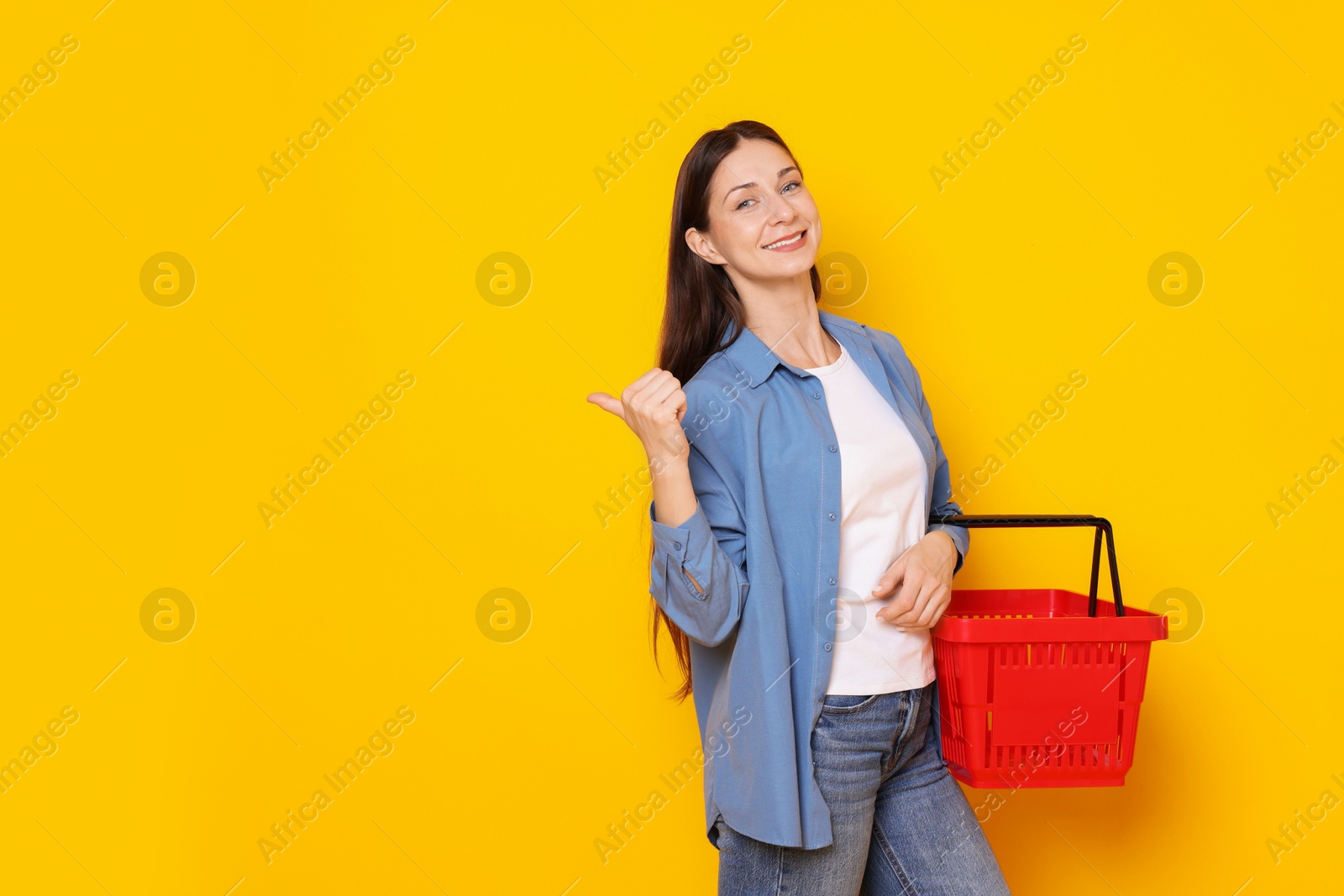 Smiling woman with shopping basket pointing at something on yellow background. Space for text Photo of Smiling woman with shopping basket pointing at something on yellow background. Space for text