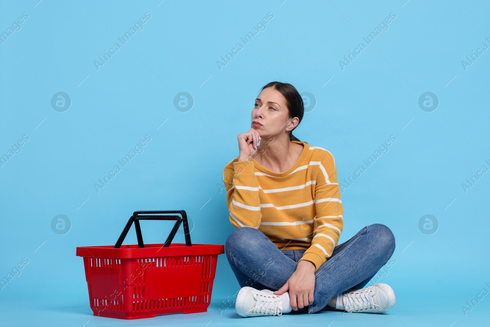 Thoughtful woman and shopping basket on light blue background Photo of Thoughtful woman and shopping basket on light blue background