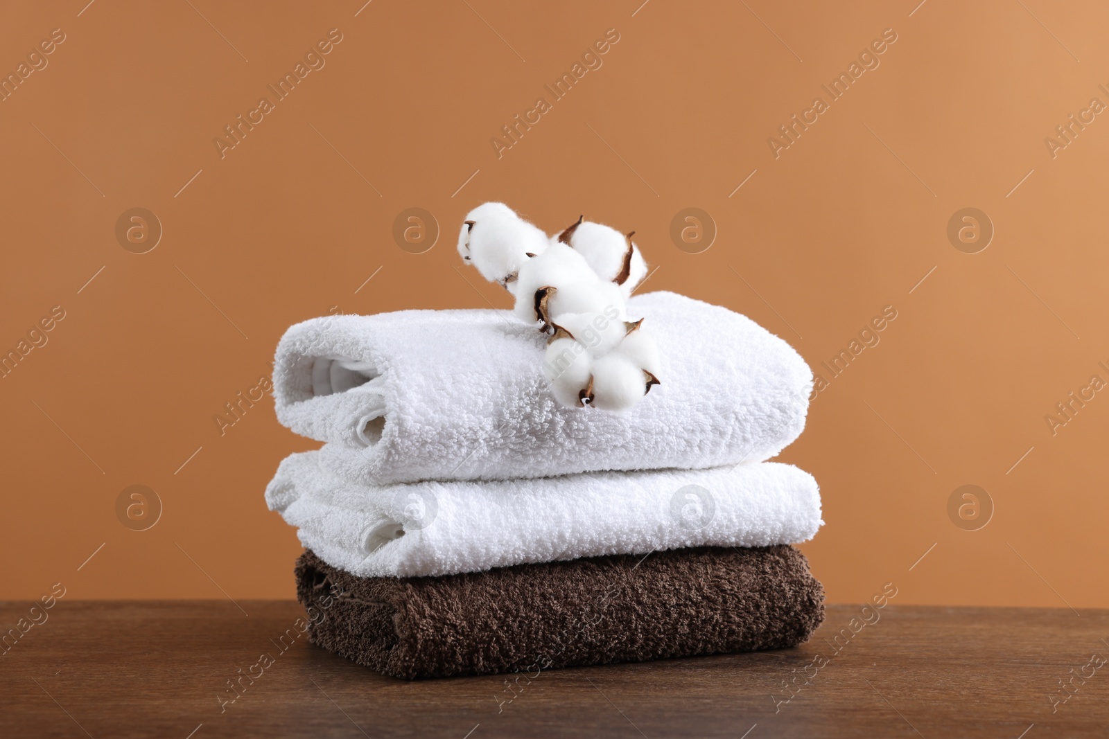 Stack of clean towels and cotton flowers on wooden table against brown background Photo of Stack of clean towels and cotton flowers on wooden table against brown background