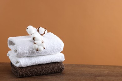 Photo of Stack of clean towels and cotton flowers on wooden table against brown background, space for text