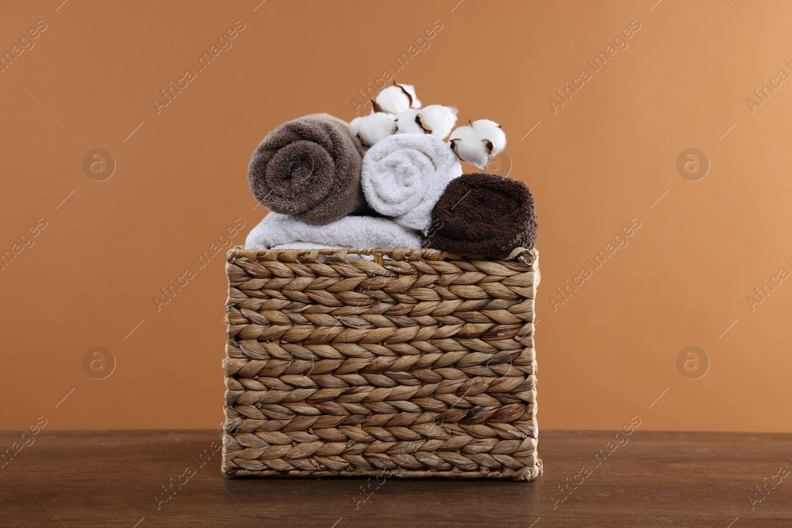 Clean towels and cotton flowers in basket on wooden table against brown background Photo of Clean towels and cotton flowers in basket on wooden table against brown background