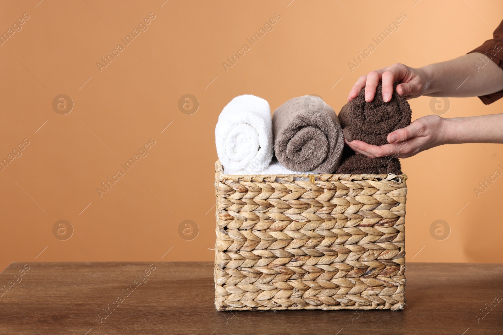 Woman taking clean towel from basket at wooden table against brown background, closeup. Space for text Photo of Woman taking clean towel from basket at wooden table against brown background, closeup. Space for text