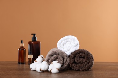 Photo of Clean terry towels, cotton flowers and cosmetic products on wooden table against brown background