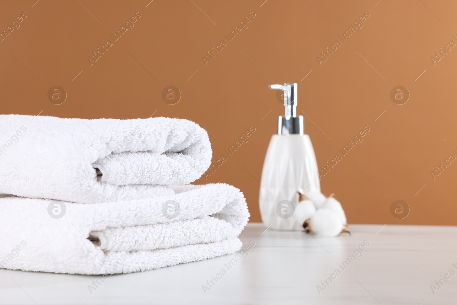 Clean terry towels, cosmetic product and cotton flower on light table against brown background Photo of Clean terry towels, cosmetic product and cotton flower on light table against brown background
