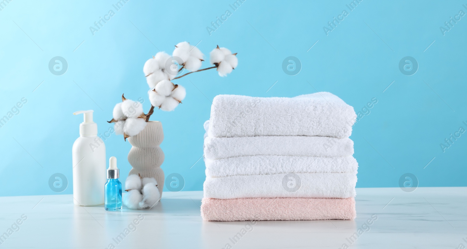 Stack of clean towels, cotton flowers and cosmetic products on white marble table against light blue background Photo of Stack of clean towels, cotton flowers and cosmetic products on white marble table against light blue background
