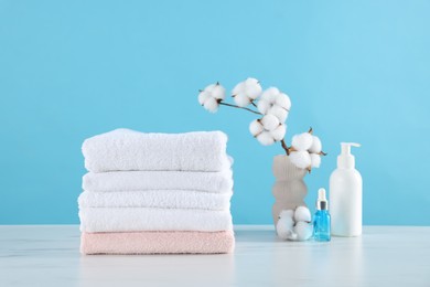 Photo of Stack of clean towels, cotton flowers and cosmetic products on white marble table against light blue background