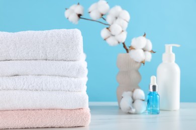Photo of Stack of clean towels, cotton flowers and cosmetic products on white marble table against light blue background, closeup