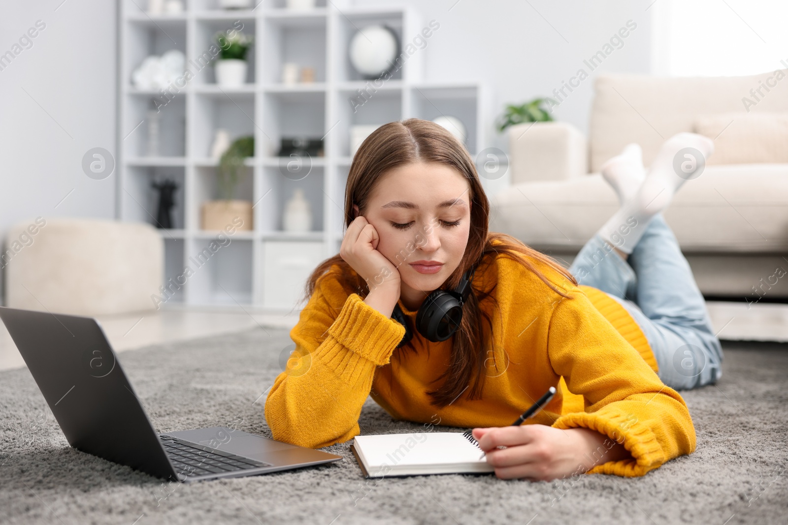 Teenage girl with headphones doing homework near laptop on floor at home Photo of Teenage girl with headphones doing homework near laptop on floor at home