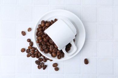 Photo of Aromatic coffee beans, cup and saucer on white tiled table, flat lay