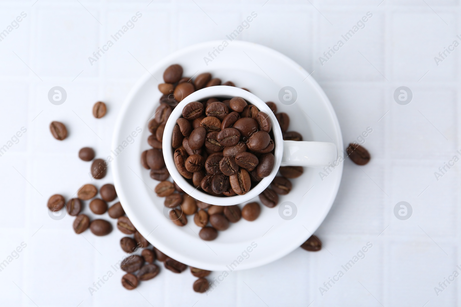 Aromatic coffee beans, cup and saucer on white tiled table, flat lay Photo of Aromatic coffee beans, cup and saucer on white tiled table, flat lay