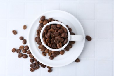 Photo of Aromatic coffee beans, cup and saucer on white tiled table, flat lay