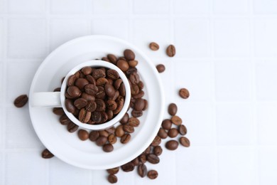 Photo of Aromatic coffee beans, cup and saucer on white tiled table, flat lay. Space for text