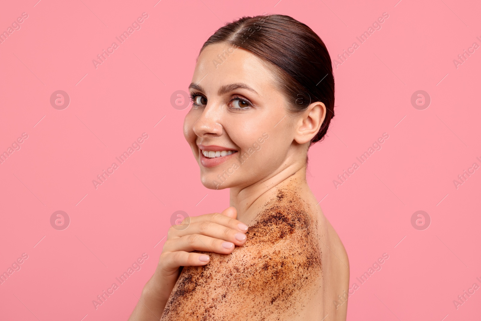 Smiling woman applying body scrub onto her shoulder against pink background Photo of Smiling woman applying body scrub onto her shoulder against pink background