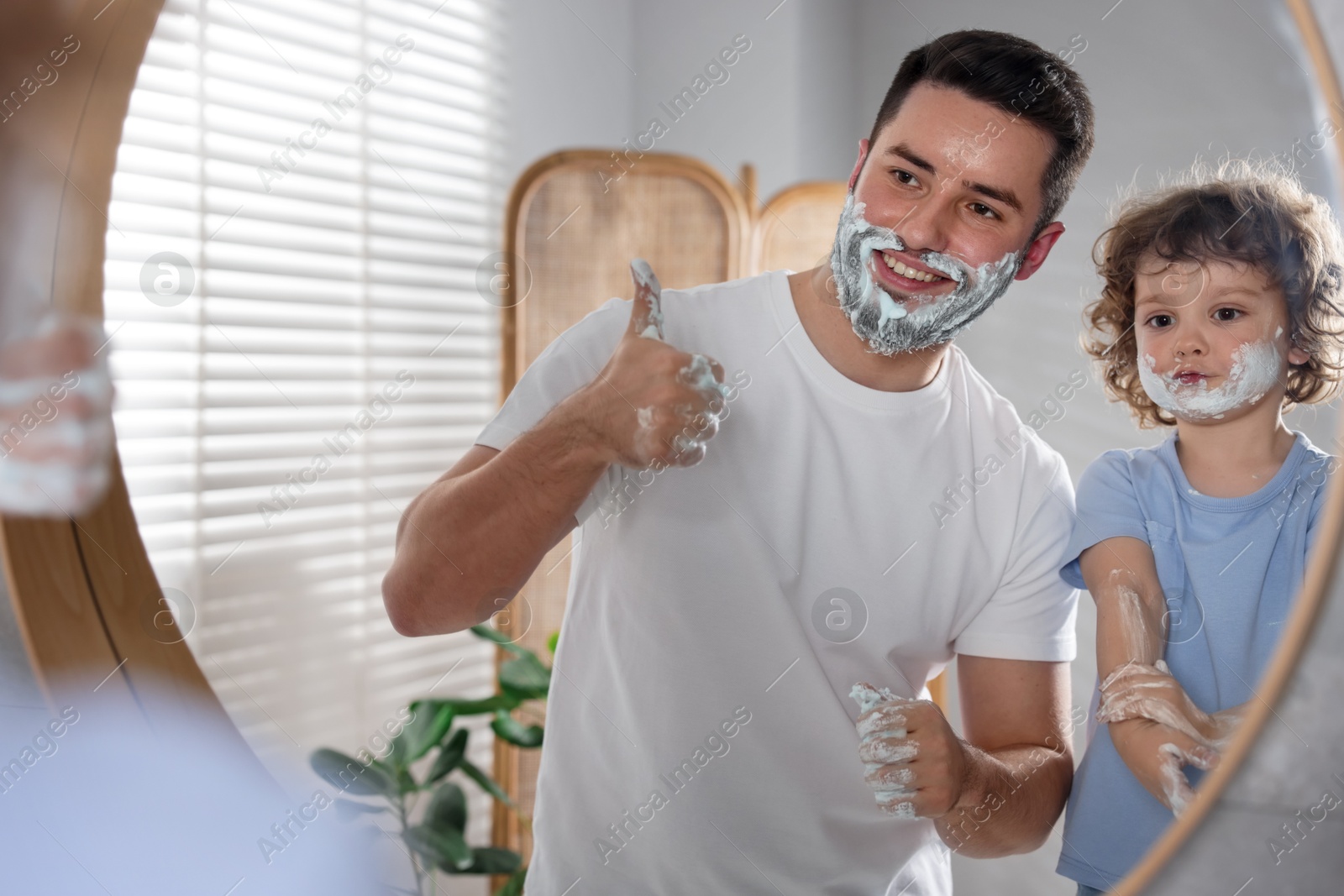Photo of Happy father and his son with shaving cream on their faces showing thumbs up near mirror in bathroom