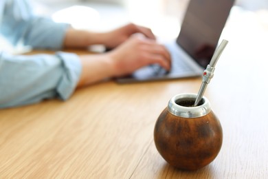 Photo of Man using laptop at wooden table indoors, focus on traditional yerba mate tea