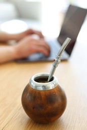 Photo of Man using laptop at wooden table indoors, focus on traditional yerba mate tea