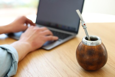 Photo of Man using laptop at wooden table indoors, focus on traditional yerba mate tea