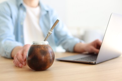 Photo of Man using laptop at wooden table indoors, focus on traditional yerba mate tea