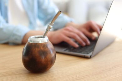 Photo of Man using laptop at wooden table indoors, focus on traditional yerba mate tea