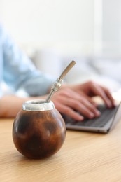 Photo of Man using laptop at wooden table indoors, focus on traditional yerba mate tea