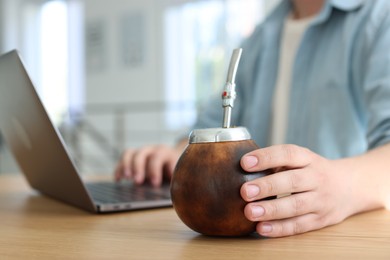 Photo of Man using laptop at wooden table indoors, focus on traditional yerba mate tea