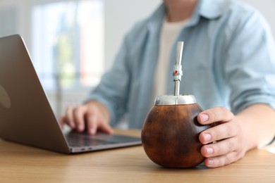 Photo of Man using laptop at wooden table indoors, focus on traditional yerba mate tea