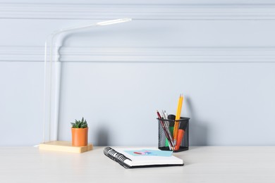 Photo of Different stationery, lamp and floral decor on office table near light wall