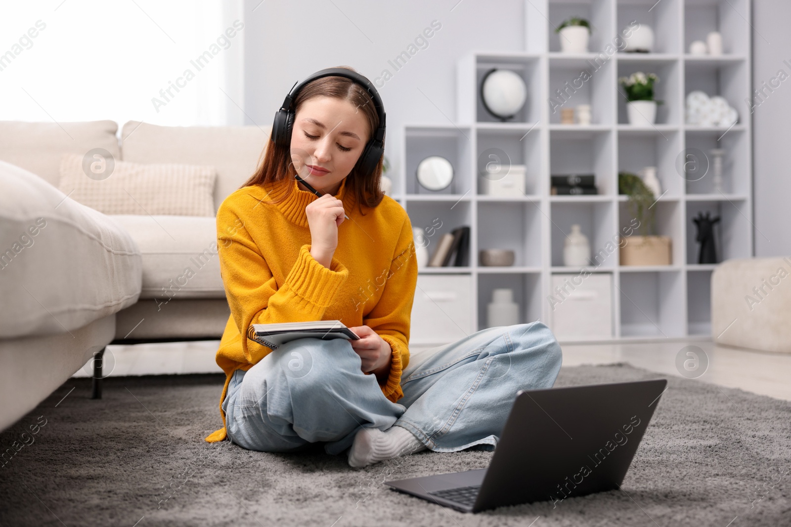 Photo of Teenage girl in headphones doing homework near laptop on floor at home