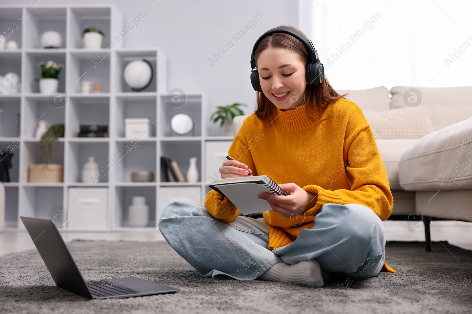 Photo of Teenage girl in headphones doing homework near laptop on floor at home