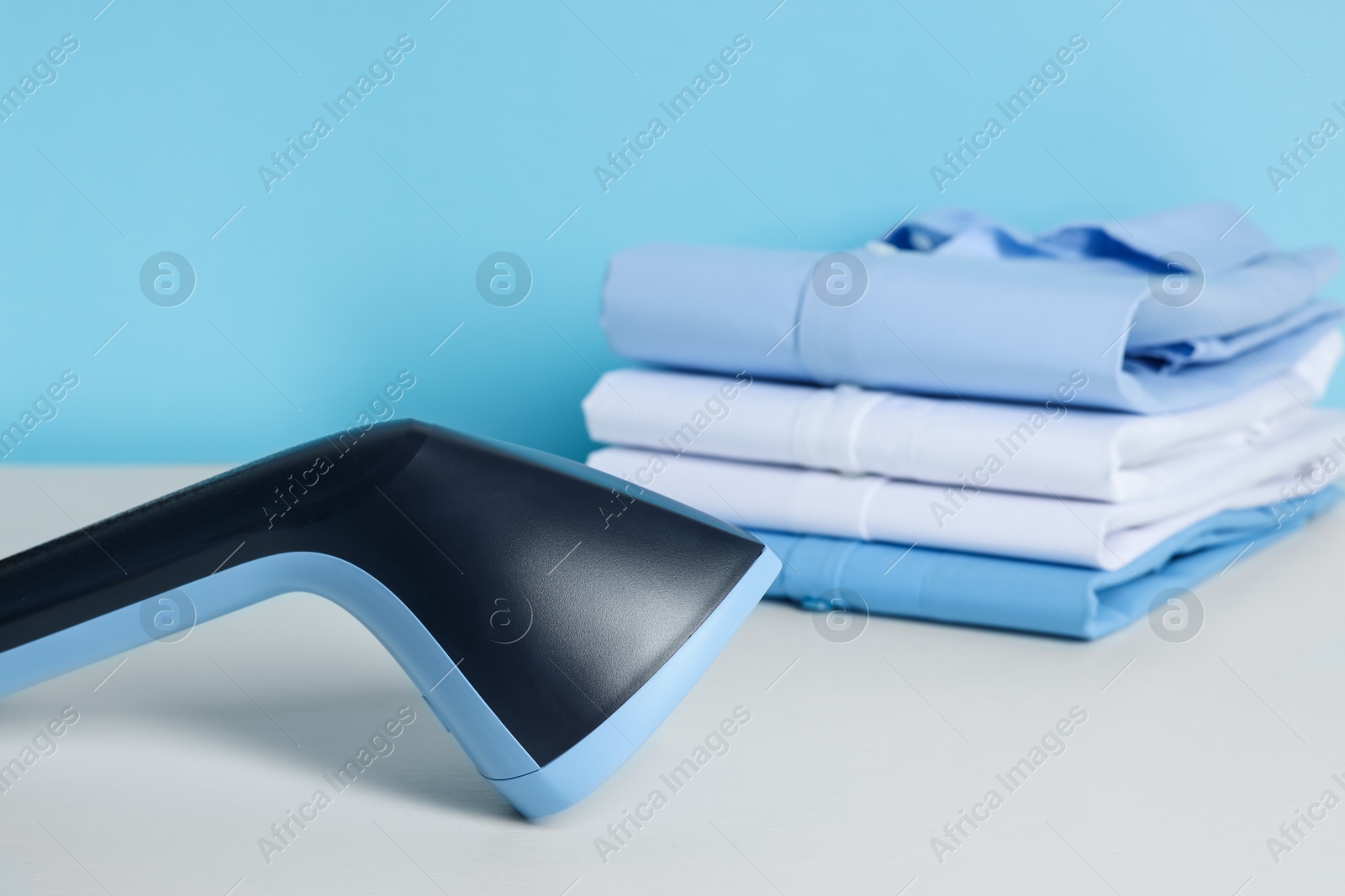 Upright steamer and stack of folded shirts on white table against light blue background, closeup Photo of Upright steamer and stack of folded shirts on white table against light blue background, closeup